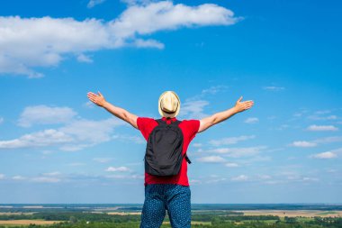 Traveler man with backpack,arms outstretched stand on blue sky white cloud summer landscape day.Man wears hat,red t-shirt blue shorts.Back view
