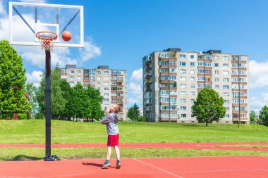 Oyun sahasında basketbol oynayan neşeli genç bir çocuk. Çocuk yaz günü oyun sahasında Red Court maçında atış yapıyor. Güneşli şehir günü..
