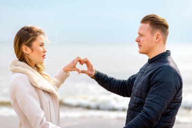 Romantik genç çift kalplerini oluşturuyor, birbirlerine bakıyor ve sahilde dururken gülümsüyorlar..