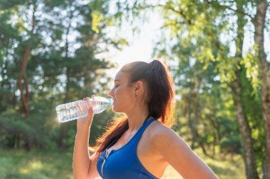 Ormanda şişeden su içen bir kız. Ormanda elinde su şişesi olan genç bir kadın portresi. Spor giyim sektöründeki pozitif kadın koşudan sonra dinleniyor. Güneşli yaz gününde