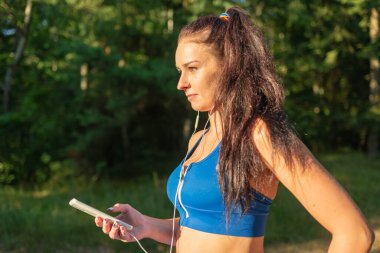 Ormanda egzersiz yapan genç ve güzel bir bayan kulaklıkla müzik dinliyor. Yaz ormanında kulaklıkla müzik dinleyen mutlu genç bir kadın. Yakından..