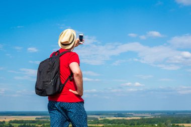 Sırt çantalı genç adam akıllı telefon kullanarak selfie çekiyor. Gökyüzü bulutlu yaz mevsimi aralığı. Manzara yaz arkaplanı. Aktif spor sırt çantalı sağlıklı yaşam tarzı konsepti..