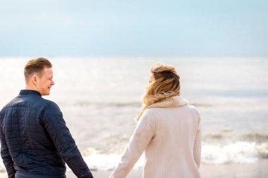 Genç aşık çift, çekici erkek ve kadın sahilde romantik bir akşam geçiriyorlar, el ele tutuşup gün batımını izliyorlar.