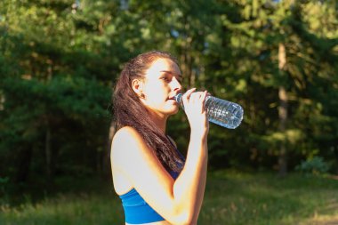 Ormanda şişeden su içen bir kız. Ormanda elinde su şişesi olan genç bir kadın portresi. Spor giyim sektöründeki pozitif kadın koşudan sonra dinleniyor. Güneşli yaz gününde
