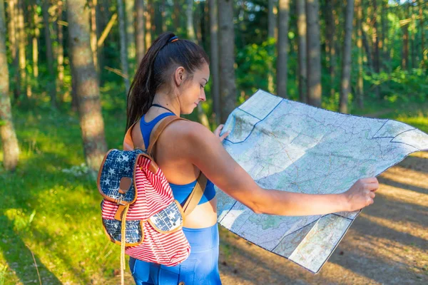 Young pretty hiker woman with backpack holding map in summer forest.female traveler checking destination on map.Young female hiker using map for orienteering in the forest.Active lifestyle concept.