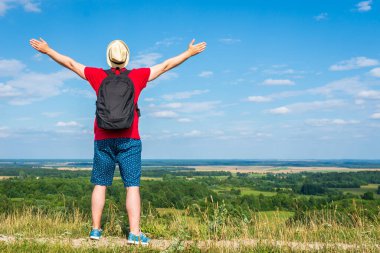 Sırt çantalı turist. Sırt çantalı, kollarını açmış gezgin. Mavi gökyüzü beyaz bulutlu yaz manzarasında duruyor.
