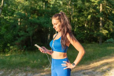 Ormanda egzersiz yapan genç ve güzel bir bayan kulaklıkla müzik dinliyor. Yaz ormanlarında kulaklıkla müzik dinleyen mutlu genç bir kadın..