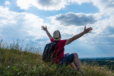 Sırt çantalı turist. Sırt çantalı, kollarını açmış gezgin. Mavi gökyüzü beyaz bulutlu yaz manzarasında duruyor.