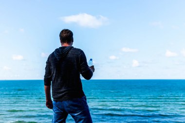 Orta yaşlı bir adam güneşli yaz günlerinde sahilde gezindikten sonra dinleniyor ve su şişesi tutuyor..