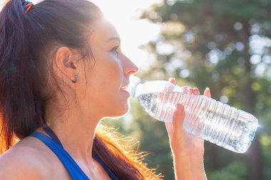 Ormanda şişeden su içen bir kız. Ormanda elinde su şişesi olan genç bir bayan. Spor giysili pozitif bayan koştuktan ve koştuktan sonra dinleniyor. Güneşli yaz gününde. Yakın çekim, yan görüş..