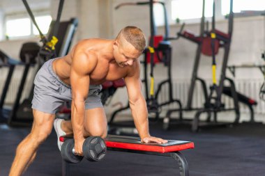 Güçlü vücut geliştirici, spor salonunda dambılla egzersiz yapıyor. Genç, yakışıklı, yetişkin bir erkek, modern spor salonunda tek kollu halterde kürek çekiyor. Boşluğu kopyala..