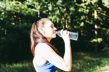 Ormanda şişeden su içen bir kız. Ormanda elinde su şişesi olan genç bir kadın portresi. Spor giyim sektöründeki pozitif kadın koşudan sonra dinleniyor. Güneşli yaz gününde