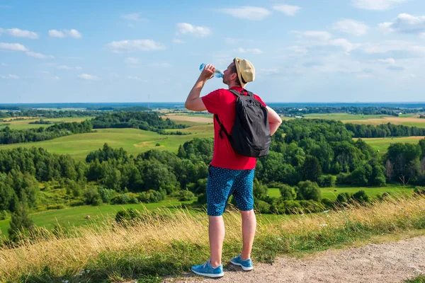 Gezgin dağlarda su içiyor. Sırt çantalı gezgin dağın tepesinde dinleniyor ve güneşli yaz gününde şişe su içiyor..