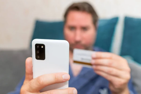 Young man buying fashion items through the Internet on cellphone by using credit card.Online Payment. Frontal portrait of man holding debit card and using smartphone for shopping at home