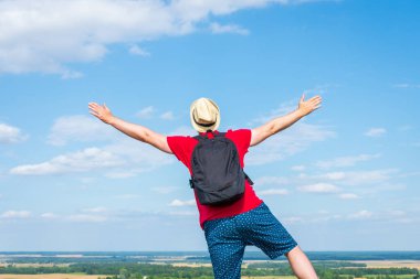 Sırt çantalı turist. Sırt çantalı, kollarını açmış gezgin. Mavi gökyüzü beyaz bulutlu yaz manzarasında duruyor.