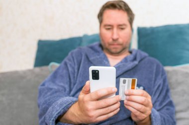 Moda ürünlerini cep telefonuyla kredi kartıyla alan genç bir adam. Online Ödeme. Bankamatik kartı tutan adamın ön portresi ve evden alışveriş için akıllı telefon kullanımı