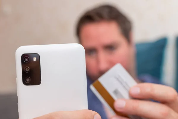 Young man buying fashion items through the Internet on cellphone by using credit card.Online Payment. Frontal portrait of man holding debit card and using smartphone for shopping at home