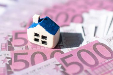 Euro banknotlarının üzerine yerleştirilmiş minyatür oyuncak evi. Gayrimenkul maliyeti, fiyatları, ev satın alma veya kiralama kavramı, hipotezi kredisi, ilgi çekici odaklanma, yakın plan. Bulanık arka plan ve ön plan.