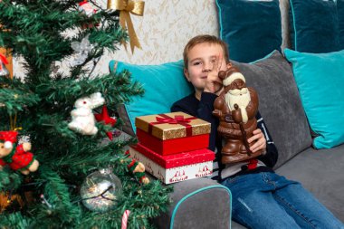 Kanepede oturan çocuk Noel Baba çikolatalı hediyeler tutuyor. Evde süslenmiş Noel ağacının yanında..