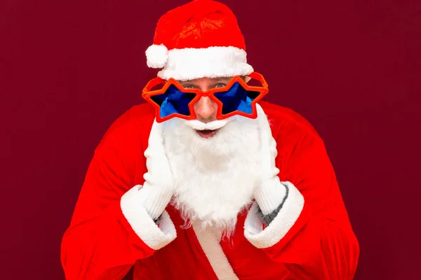 身穿礼服头戴星形眼镜的圣诞老人兴奋的白胡子张大了嘴凝视着眼睛听着令人难以置信的消息在红工作室的背景上被隔离了 — 图库照片