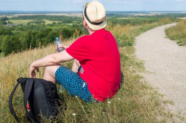 Gezgin dağlarda su içiyor. Sırt çantalı gezgin dağın tepesinde dinleniyor ve güneşli yaz gününde şişe su içiyor..