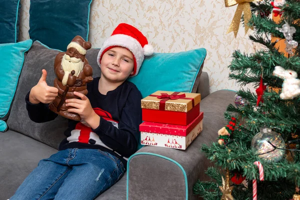 Kanepede oturan çocuk, elinde Noel Baba çikolatası ile baş parmağını kaldırıyor..