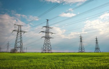 Güneşli bir günde mavi gökyüzünde bulutlar olan düz ve yeşil tarım arazisindeki yüksek voltaj hatları ve elektrik direkleri. Bulutlu ve yağmurlu. Buğday büyüyor.