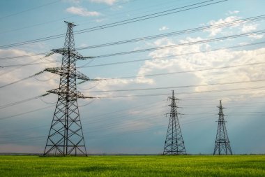Güneşli bir günde mavi gökyüzünde bulutlar olan düz ve yeşil tarım arazisindeki yüksek voltaj hatları ve elektrik direkleri. Bulutlu ve yağmurlu. Buğday büyüyor.
