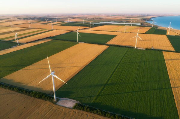 Aerial view of renewable windmills turbines supplying cultivation area with eco power getting energy from wind blowing on vast area of agriculture meadows next to sea. Alternative electricity