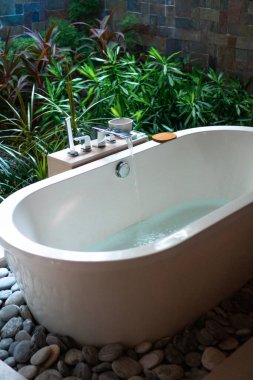 Butik tatil köyünün yemyeşil bahçesinde, akan suyu olan, modern, serbest beyaz bir küvet. Tropikal titreşimler ve tertemiz rahatlama atmosferi..