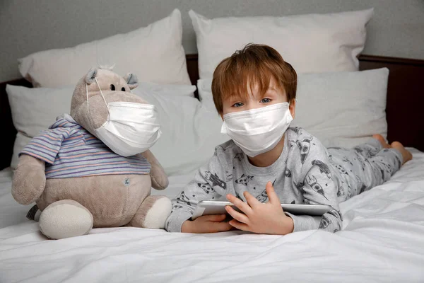 Niño enfermo de cuatro años con una máscara de protección médica utiliza una tableta mientras está acostado en una cama en casa. el concepto de cuarentena y epidemia — Foto de Stock