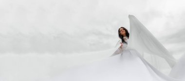 Uzun beyaz elbiseli güzel bir kadın bulutların karşısındaki bir meleğe benziyor. Elbise rüzgar yüzünden gelişiyor.