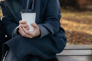 Yakın plan - Parkta tek kullanımlık kahve fincanı tutan, ceketli beyaz bir kadının elleri
