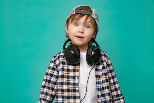 Cute 4-5 year old boy in shirt and baseball cap listening to music with headphones on isolated green background — Stock Photo, Image