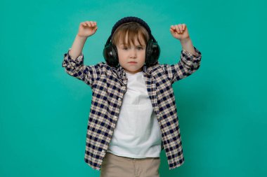 Küçük, şık bir çocuk, 4-5 yaşında, kareli tişörtlü ve kafasında şapkası olan beyaz bir çocuk ellerini yana açmış, bir çocuğun pastel mavi arka planına karşı durarak dans ediyor.