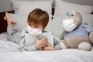 Hasta 5 yaşında, koruyucu maskeli bir çocuk evde yatakta yatarken tablet bilgisayar kullanıyor. Karantina ve salgın koronavirüs türü kavramı.