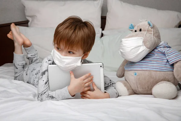 Niño enfermo de cinco años con una máscara de protección médica utiliza una tableta mientras está acostado en una cama en casa. concepto de cuarentena y cepa del coronavirus epidémico. — Foto de Stock