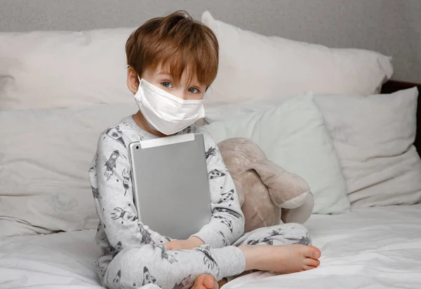 Hermoso niño de cinco años de edad, en una máscara se sienta en la cama y sostiene una tableta en sus manos. Conceptos de la cepa coronovirus — Foto de Stock