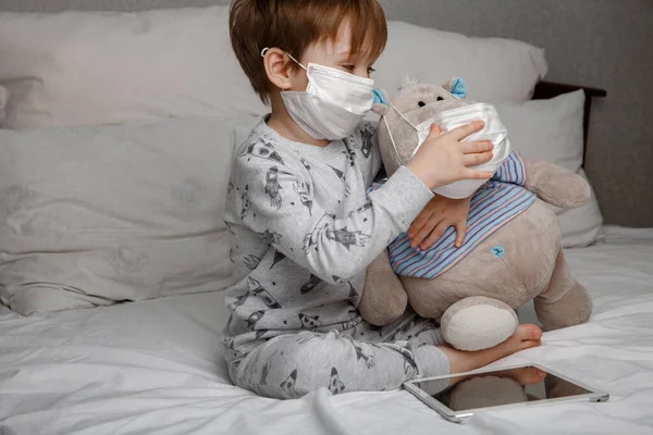 Niño enfermo de cinco años con una máscara médica protectora juega con su juguete favorito mientras está acostado en una cama en casa. Concepto de cuarentena y cepa epidémica del coronavirus. — Foto de Stock