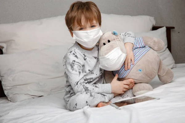 Niño enfermo de cinco años con una máscara médica protectora juega con su juguete favorito mientras está acostado en una cama en casa. Concepto de cuarentena y cepa epidémica del coronavirus. — Foto de Stock