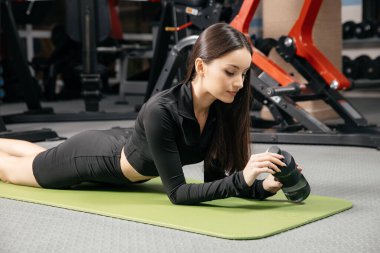 Güzel esmer kadın spor salonunun paspasına uzanıyor ve sıkı bir çalışmadan sonra su içiyor. Ara verme kavramı