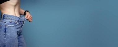 30 'larında, geniş kot pantolon ve beyaz gömlek giyen çekici, narin genç bir kadın başarılı bir kilo kaybı gösteriyor. Diyet ve kilo kaybı konsepti..