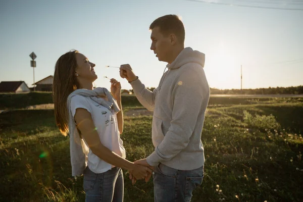 Hermosa pareja blanca joven de unos treinta años, divirtiéndose en el pueblo al aire libre, soplándose dientes de león el uno al otro y sonriendo dulcemente, espacio libre — Foto de Stock