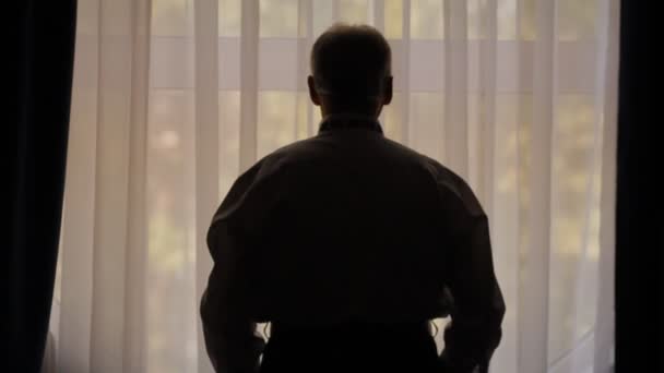 Старик в белой рубашке смотрит в окно — стоковое видео