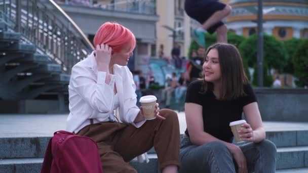 Две девушки разговаривают и смеются сидя на гранитной лестнице и пьют кофе — стоковое видео