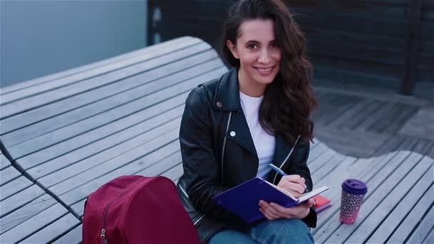 Vrouwelijke studente die in notebook buiten op de moderne houten bank zit te schrijven — Stockvideo
