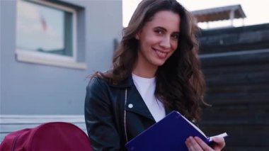 Defterle yazan kız öğrenci dışarıda, modern ahşap bankta oturuyor.