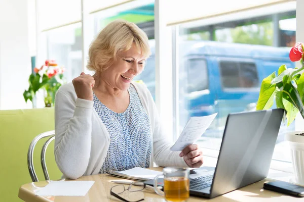 A woman in the office, cafe with a laptop working at a table in the room. A middle-aged woman enjoys success