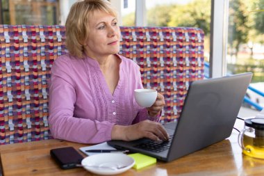 Dizüstü bilgisayarı olan bir kadın odadaki masada çalışıyor. Orta yaşlı bir kadın internette bilgisayarla iletişim kuruyor. Çay içiyor.