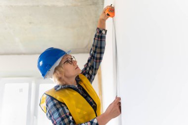 Kadın bir inşaat işçisi yeni bir binayı teftiş ediyor. Sarı yelek giyen orta yaşlı bir kadın ölçü alıyor..